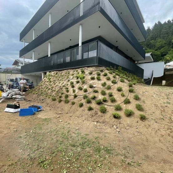 Ein modernes Gebäude mit einem geneigten Dach und mehreren Balkonen, das auf einem Hügel mit Begrünung liegt. Eine Person steht in der Nähe eines Autos. Verschiedene Gegenstände liegen auf dem Boden, und es gibt Bäume im Hintergrund.