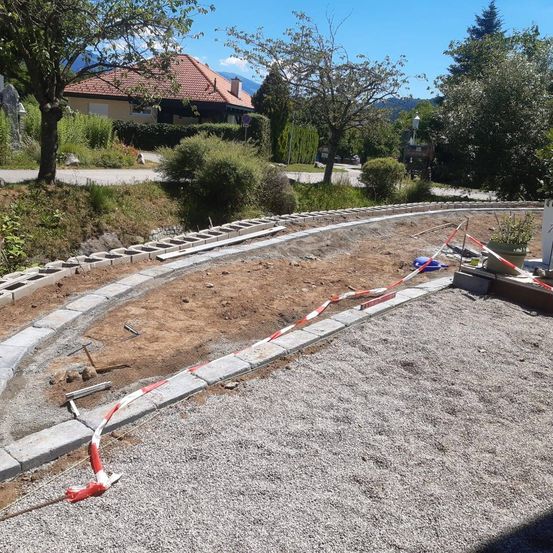 Eine Baustelle mit Kies und Blöcken, ein Hammer und Absperrband, ein Haus und Bäume im Hintergrund.