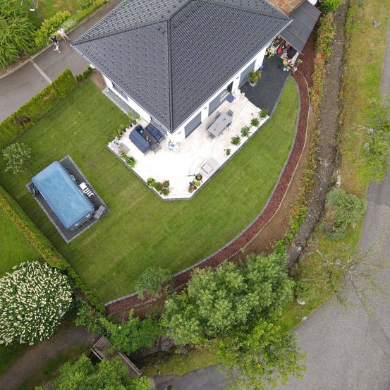 Ein Luftbild eines modernen Hauses mit grünem Rasen und Terrasse, mit Swimmingpool, Garten und mehreren Bäumen.