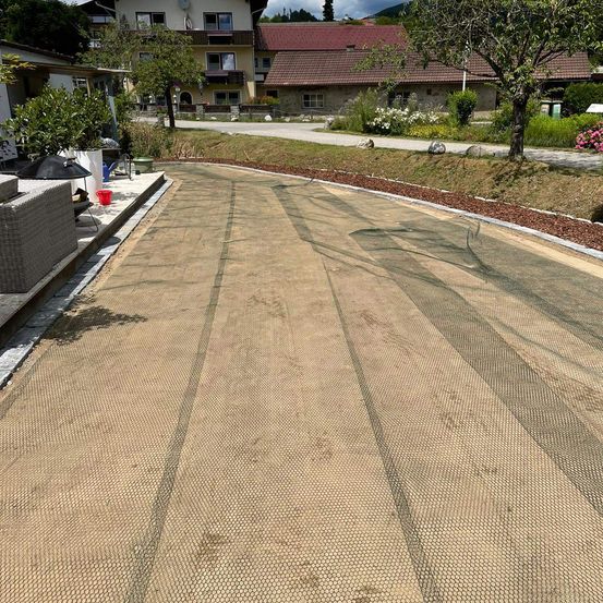 Eine leere Einfahrt ist mit einem Schutznetz bedeckt. Im Hintergrund sind Häuser und Bäume zu sehen. Ein roter Eimer steht neben der Einfahrt.