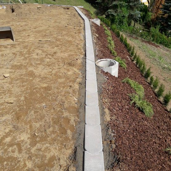 Eine Baustelle im Freien zeigt einen Betonrand, einen Erdhaufen und einen Entwässerungsgraben.