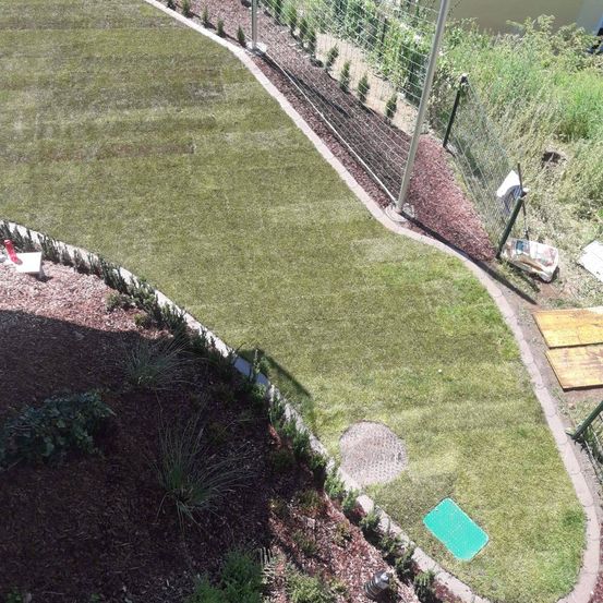 Ein Vogelperspektive auf einen gut gepflegten Rasen, der von Kies und Pflanzen gesäumt wird, mit einem eingezäunten Bereich und einem hölzernen Brett in der Nähe.