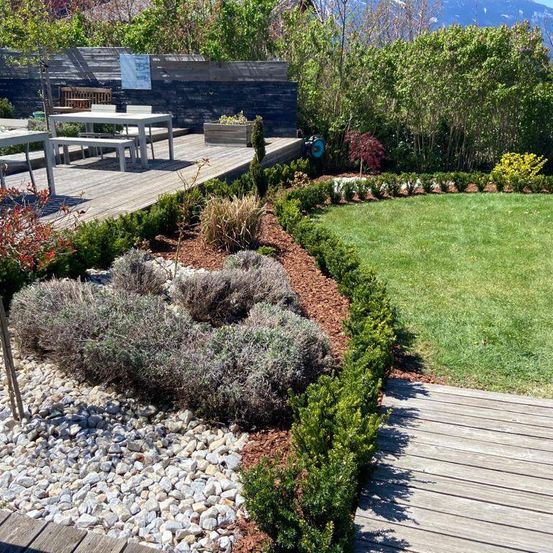 Ein Garten mit einer Holzterrasse, Tisch und Bank. Der Rasen wird von Sträuchern und Kräutern gesäumt. Berge sind in der Ferne sichtbar.