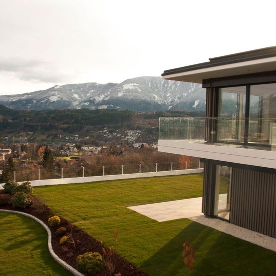 Luftaufnahme eines modernen Hauses mit Balkon, das einen üppigen grünen Rasen und einen entfernten verschneiten Berg zeigt.