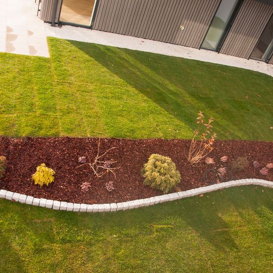 Ein Blick von oben auf einen grünen Rasen mit einem geschwungenen Gartenbeet, das mit brauner Rinde und kleinen Pflanzen gesäumt ist. Das Gartenbeet ist von weißen Steinen gesäumt. Dahinter steht ein Gebäude mit Glaswänden und einem hölzernen Eingang.