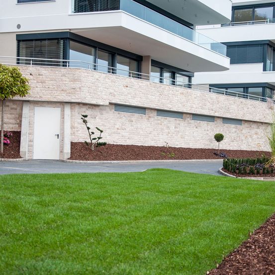 Ein modernes Gebäude mit Glasfenstern und Balkonen, umgeben von einem gepflegten Rasen mit Sträuchern und einem Baum.