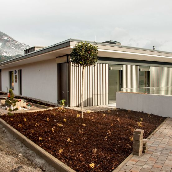 Ein modernes weißes Haus mit flachem Dach und vertikalen Streifen, vor einem Berg stehend, mit einem kleinen Garten und einem Baum in der Mitte.