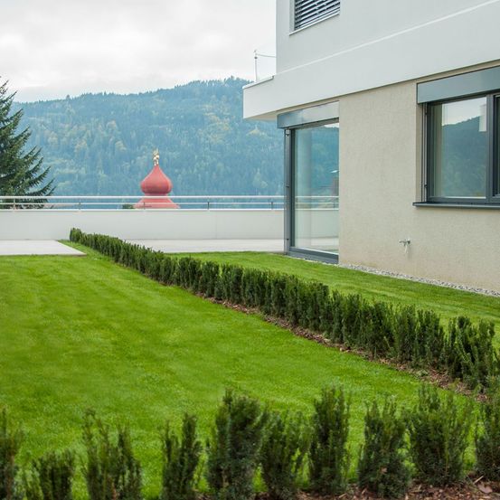 Ein modernes Haus mit grünem Rasen und Hecken, einer Glastür und einem Balkon mit Geländer, der einen roten Dom in der Ferne und eine Bergkette überblickt.
