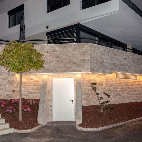 Bei Nacht ein modernes Haus mit Balkon und weißer Tür im Erdgeschoss. Der Eingang ist mit warmen Lichtern beleuchtet.