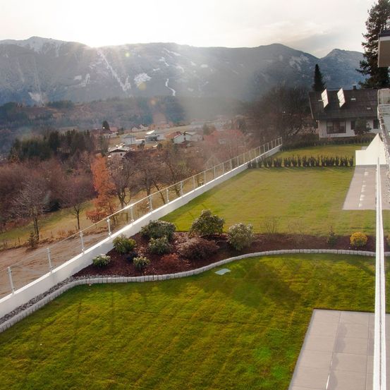 Ein Blick von oben auf einen gepflegten Rasen mit einer weißen Mauer, umgeben von Bäumen und Büschen, mit einem Haus und Bergen im Hintergrund.