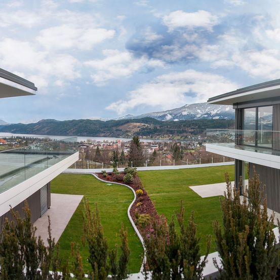 Luftaufnahme von zwei modernen Villen mit Glasbalkonen, mit Blick auf einen Rasen, einen See und verschneite Berge.