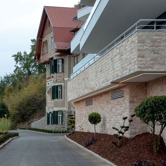 Ein modernes Gebäude mit einem roten Dach und Steinwänden steht neben einer leeren Straße, flankiert von gepflegten Hecken und kleinen Bäumen.