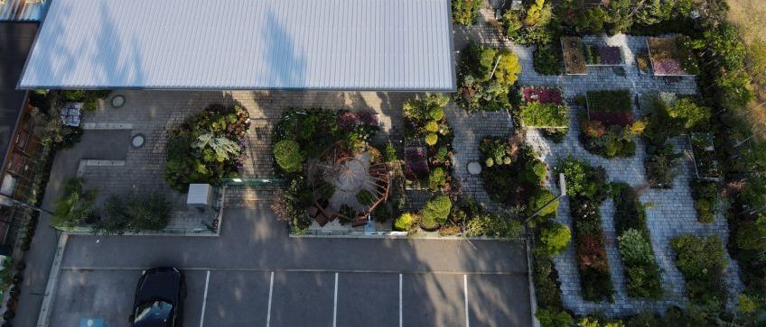 Luftaufnahme eines Gartencenters mit einem großen Gebäude, einem geparkten Auto und verschiedenen Pflanzen im Vordergrund.