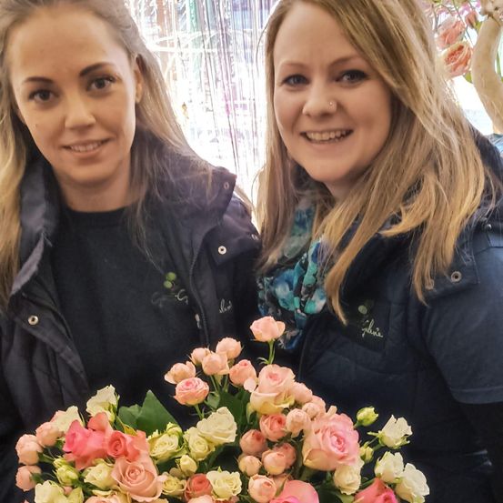 Zwei Frauen mit blonden Haaren stehen nebeneinander und lächeln. Sie halten einen Strauß rosa und weißer Rosen. Sie tragen schwarze Jacken. Hinter ihnen ist ein weißer Vorhang mit einer verschwommenen Ansicht eines Gebäudes.