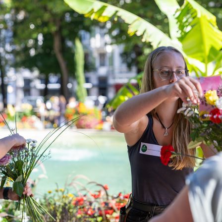 Eine Frau in einem ärmellosen Shirt mit Namensschild arrangiert Blumen in einem Garten. Sie hält eine rosa Blume in der Hand und eine andere Person hält einen Blumenstrauß. Ein Gebäude ist im Hintergrund.