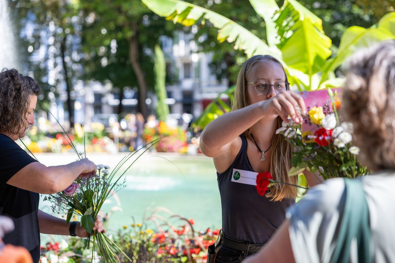 Eine Frau in einem ärmellosen Shirt mit Namensschild arrangiert Blumen in einem Garten. Sie hält eine rosa Blume in der Hand und eine andere Person hält einen Blumenstrauß. Ein Gebäude ist im Hintergrund.