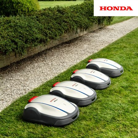 Vier graue und weiße Honda Mimo Roboter-Rasenmäher stehen auf einem Rasen neben einem Kiesweg aufgereiht. Dahinter befindet sich eine Hecke.