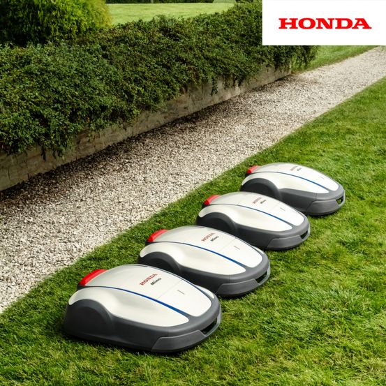 Vier graue und weiße Honda Mimo Roboter-Rasenmäher stehen auf einem Rasen neben einem Kiesweg aufgereiht. Dahinter befindet sich eine Hecke.