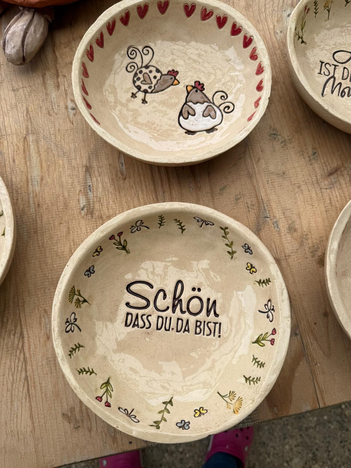 A collection of ceramic plates with floral and chicken designs, one plate has the text 'Schon, dass du da bist!' written in German.