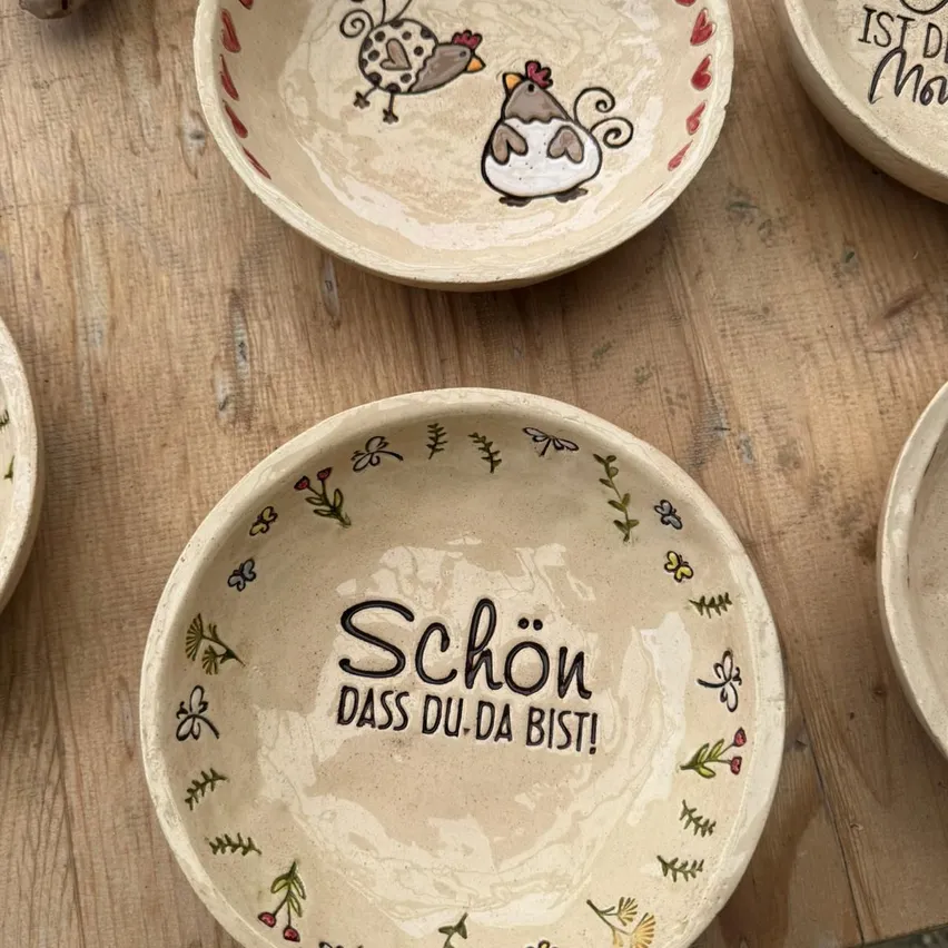 Eine Sammlung von Keramiktellern mit Blumen- und Hühnerdesigns, ein Teller hat den Text 'Schon, dass du da bist!' auf Deutsch geschrieben.