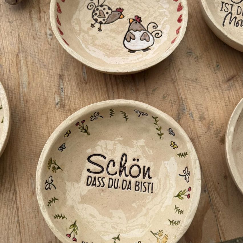Eine Sammlung von Keramiktellern mit Blumen- und Hühnerdesigns, ein Teller hat den Text 'Schon, dass du da bist!' auf Deutsch geschrieben.