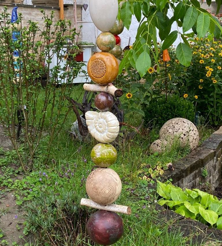 A garden display with a tall stack of colorful, spherical objects, including a pumpkin, a snail shell, and various other shapes, set against a backdrop of plants and flowers.