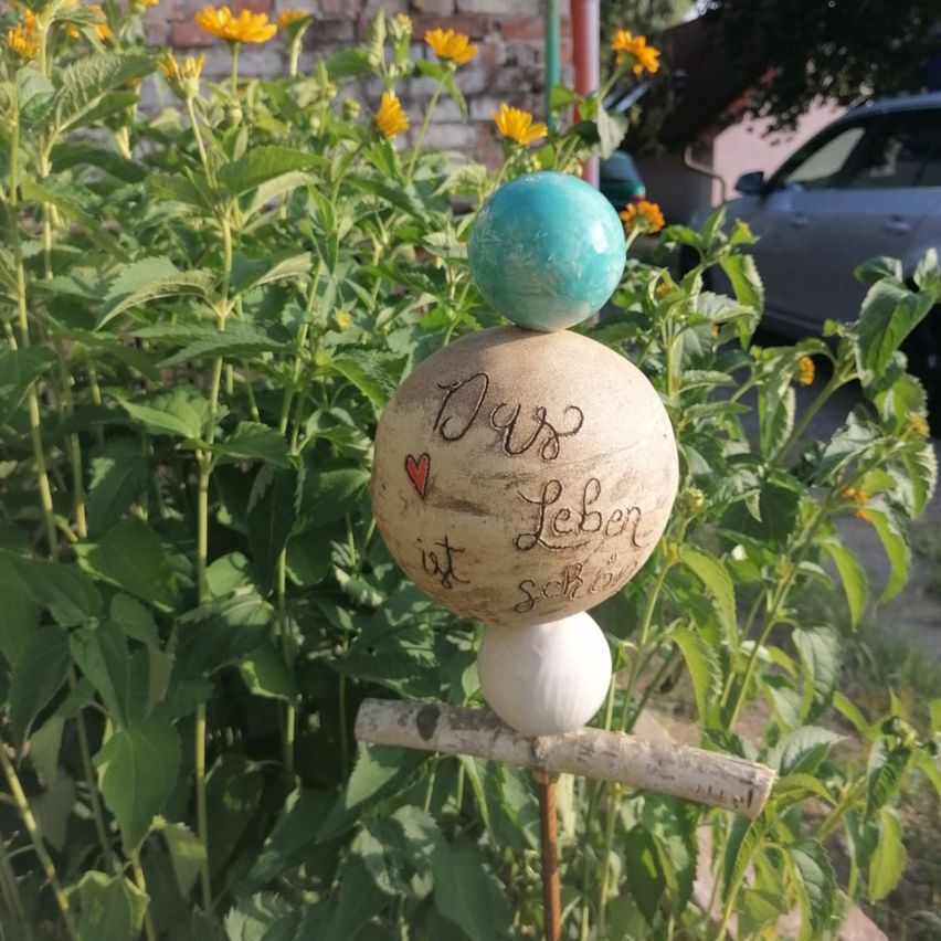 A decorative sphere with the words 'Uns ist Leben schön' written on it, placed on a stick in a garden with yellow flowers.
