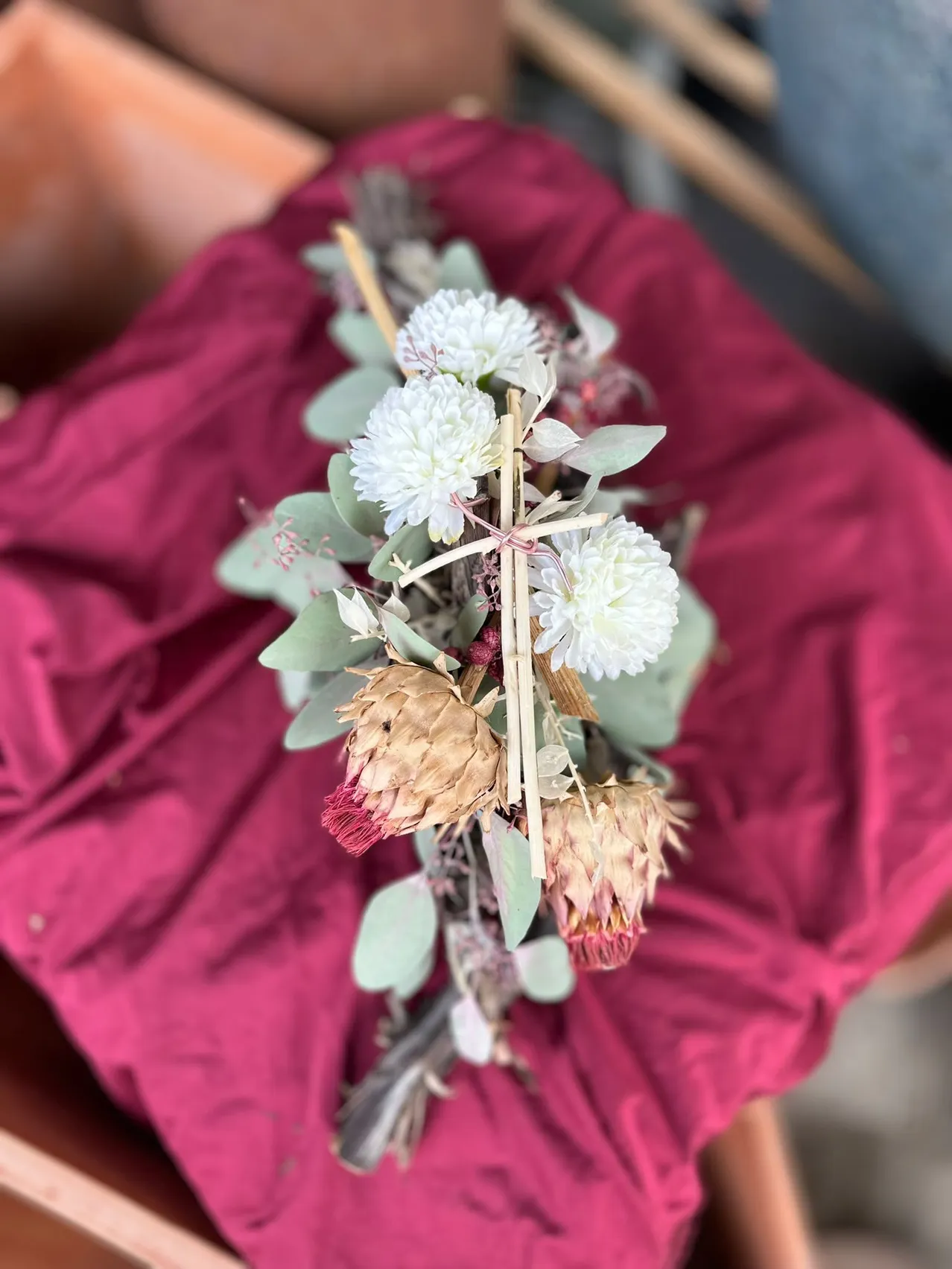 Ein Nahaufnahme einer Blumenarrangement mit weißen Blumen, grünen Blättern und einem kreuzförmigen Holzstab auf einem roten Tuch.