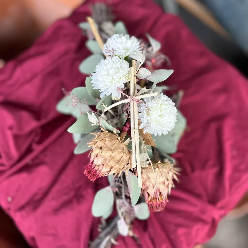 Ein Nahaufnahme einer Blumenarrangement mit weißen Blumen, grünen Blättern und einem kreuzförmigen Holzstab auf einem roten Tuch.