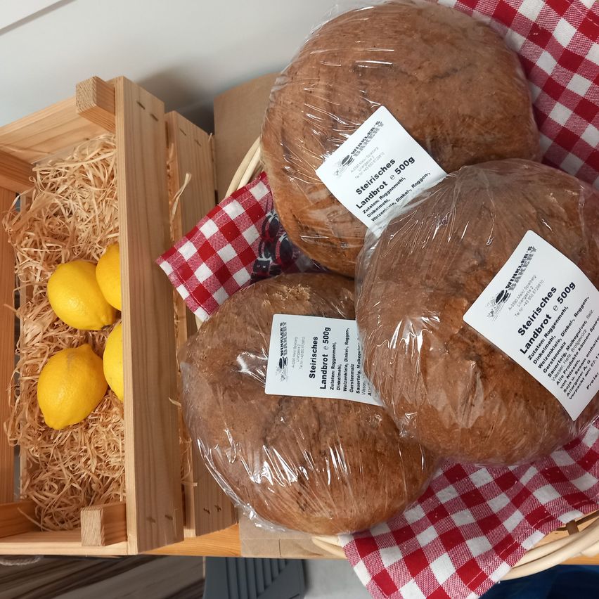 Four loaves of bread in plastic, labeled 'Landbrot 500g,' rest on a red and white cloth. Beside them is a wooden box containing lemons.