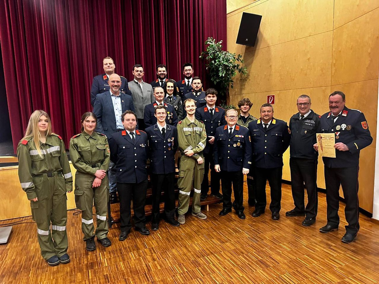 A group of uniformed individuals, some in military attire and others in suits, stand together in front of a red curtain. They appear to be posing for a photograph.