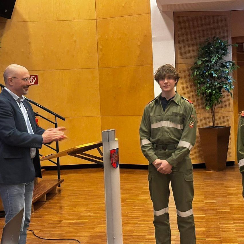 A man in a suit speaks at a podium while a young man in a military uniform stands next to a pole with a logo. A potted plant is in the background.
