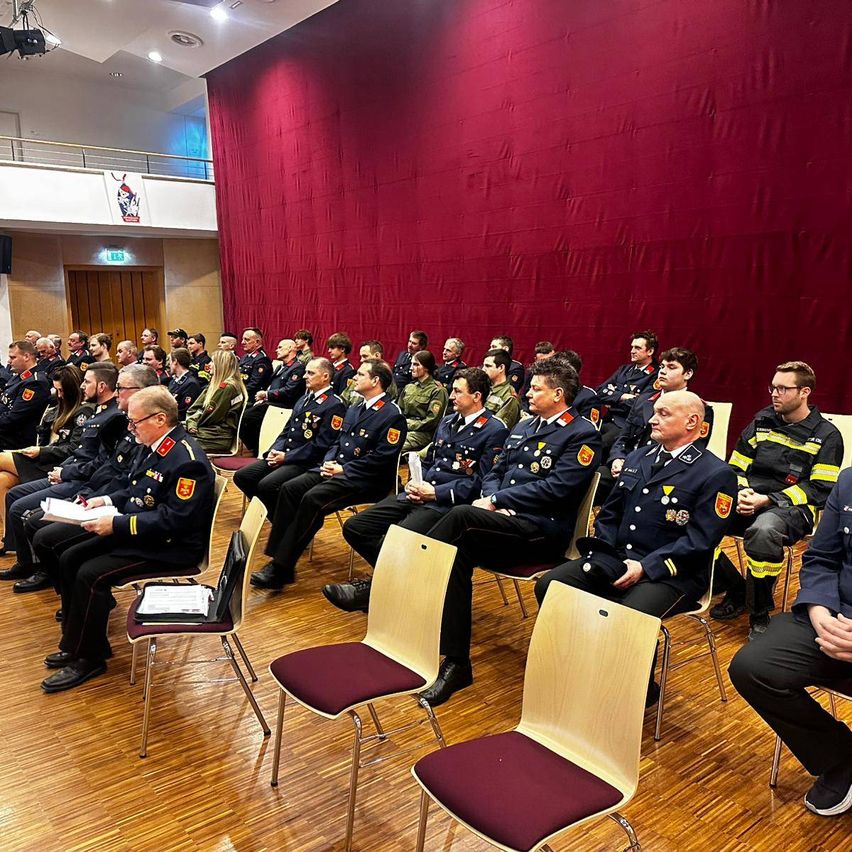 A group of uniformed individuals, possibly military, are seated in a room. Some are engaged in reading papers. The room has a red curtain and wooden flooring.