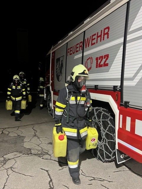 Ein Feuerwehrmann mit gelbem Helm trägt zwei gelbe Behälter und geht auf einen Feuerwehrwagen mit der Nummer 122 zu.