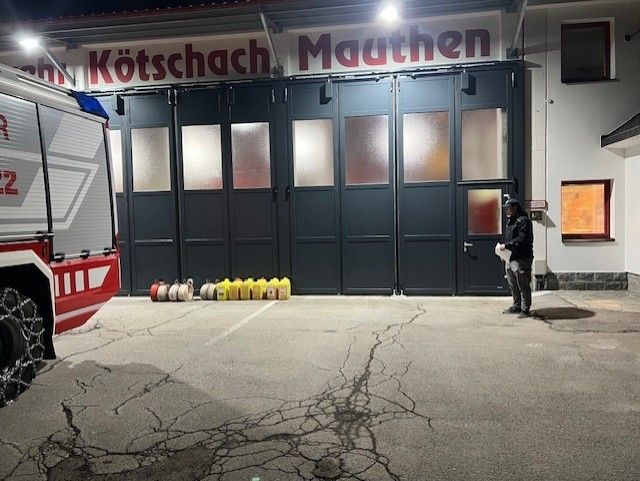 Ein Feuerwehrauto steht vor einer Garage mit einem Schild, auf dem Kotschach Mauthen steht. Eine Person steht neben gelben Containern.