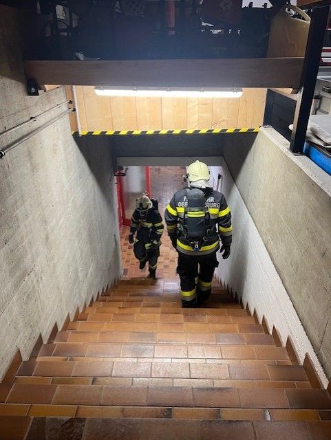 Zwei Feuerwehrleute steigen eine Ziegeltreppe hinunter, einer führt, während der andere folgt. Sie sind in voller Schutzausrüstung und Helmen gekleidet.
