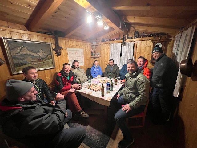 Eine Gruppe von Menschen sitzt in einer Holzhütte um einen Tisch herum, trägt Jacken und Mützen. Flaschen und Essen sind auf dem Tisch. Ein gerahmtes Bild hängt an der Wand.