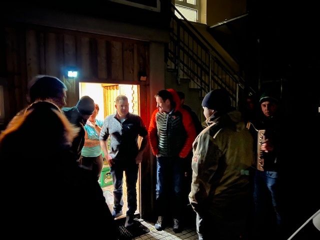 Eine Gruppe von Menschen steht in einem schwach beleuchteten Raum, möglicherweise einer Bar, mit einem Mann in der Mitte, der eine Bierflasche hält. Der Raum hat eine Holzwand, eine Treppe und ein beleuchtetes Schild.