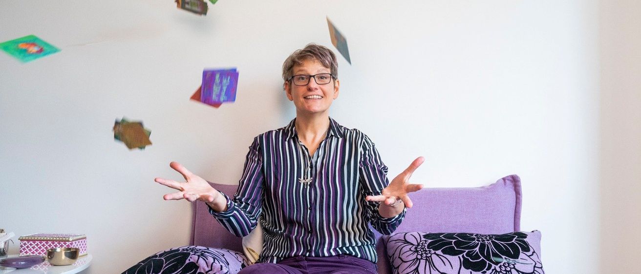 Eine Frau mit Brille sitzt auf einem lila Sofa, trägt ein gestreiftes Hemd und lila Hose. Bunte Karten schweben um sie herum, und sie hält eine Karte in der Hand.