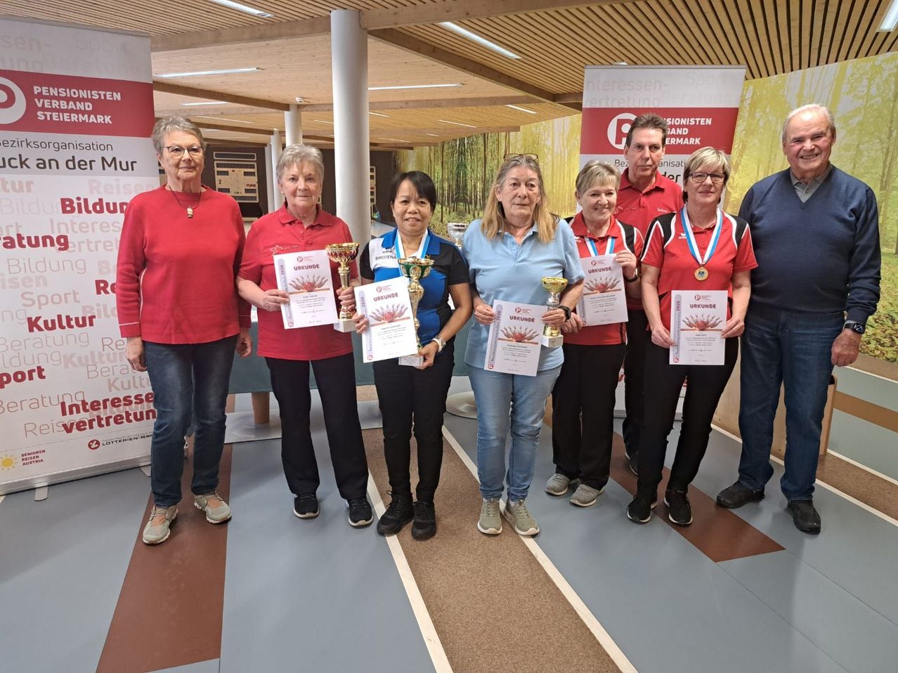 Sechs Frauen stehen in einer Reihe, halten Urkunden und Trophäen. Sie scheinen einen Sieg zu feiern. Hinter ihnen ist eine Wand mit Werbung.