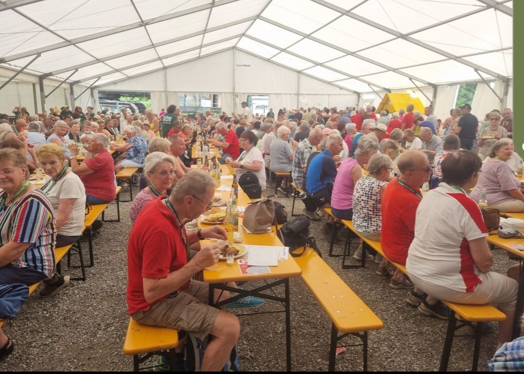Eine große Gruppe von Menschen sitzt auf Bänken in einem Zelt und isst. Das Zelt ist gut beleuchtet, mit Lampen an der Decke. Die Menschen tragen Freizeitkleidung. Einige tragen Brillen. Ein Tisch mit Essen und einer Tasche steht vor ihnen.