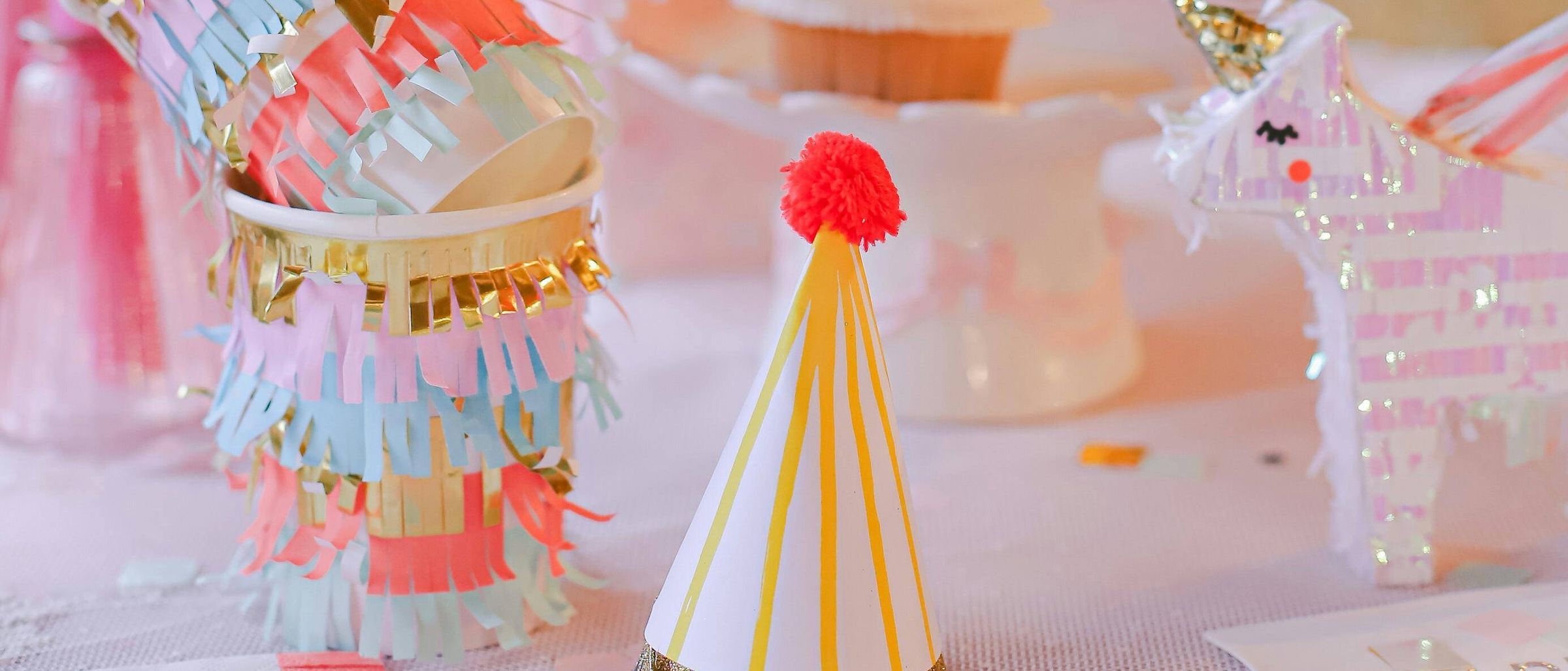 Ein festliches Geburtstags-Setup mit bunten Dekorationen, einem Cupcake und einer Partymütze. Es gibt goldverzierte Becher, ein rosa Einhorn und eine Kerze.