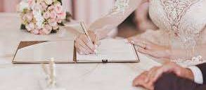 Eine Braut in einem Hochzeitskleid schreibt auf einer Trauungsurkunde am Tisch mit einem Blumenstrauß. Sie hält einen Stift in der rechten Hand.