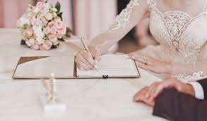 Eine Braut in einem Hochzeitskleid schreibt auf einer Trauungsurkunde am Tisch mit einem Blumenstrauß. Sie hält einen Stift in der rechten Hand.