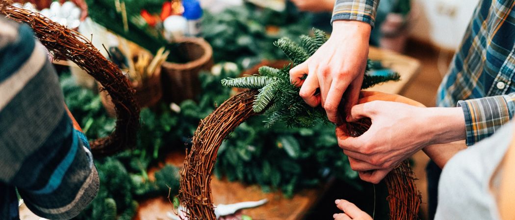 Zwei Personen sind dabei zu sehen, wie sie einen Kranz herstellen, wobei sie grünes Laub und Zweige verwenden. Eine Hand befestigt das Laub, während die andere Hand den Kranz hält. Verschiedene Weihnachtsdekorationen sind im Hintergrund zu sehen.