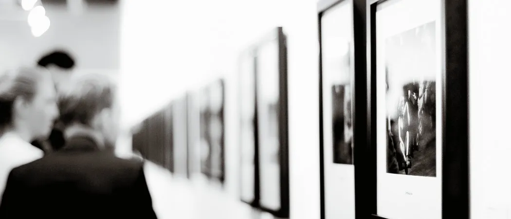 Ein schwarz-weißes Bild einer Person in einem Anzug, die gerahmte Bilder in einer Kunstgalerie betrachtet.