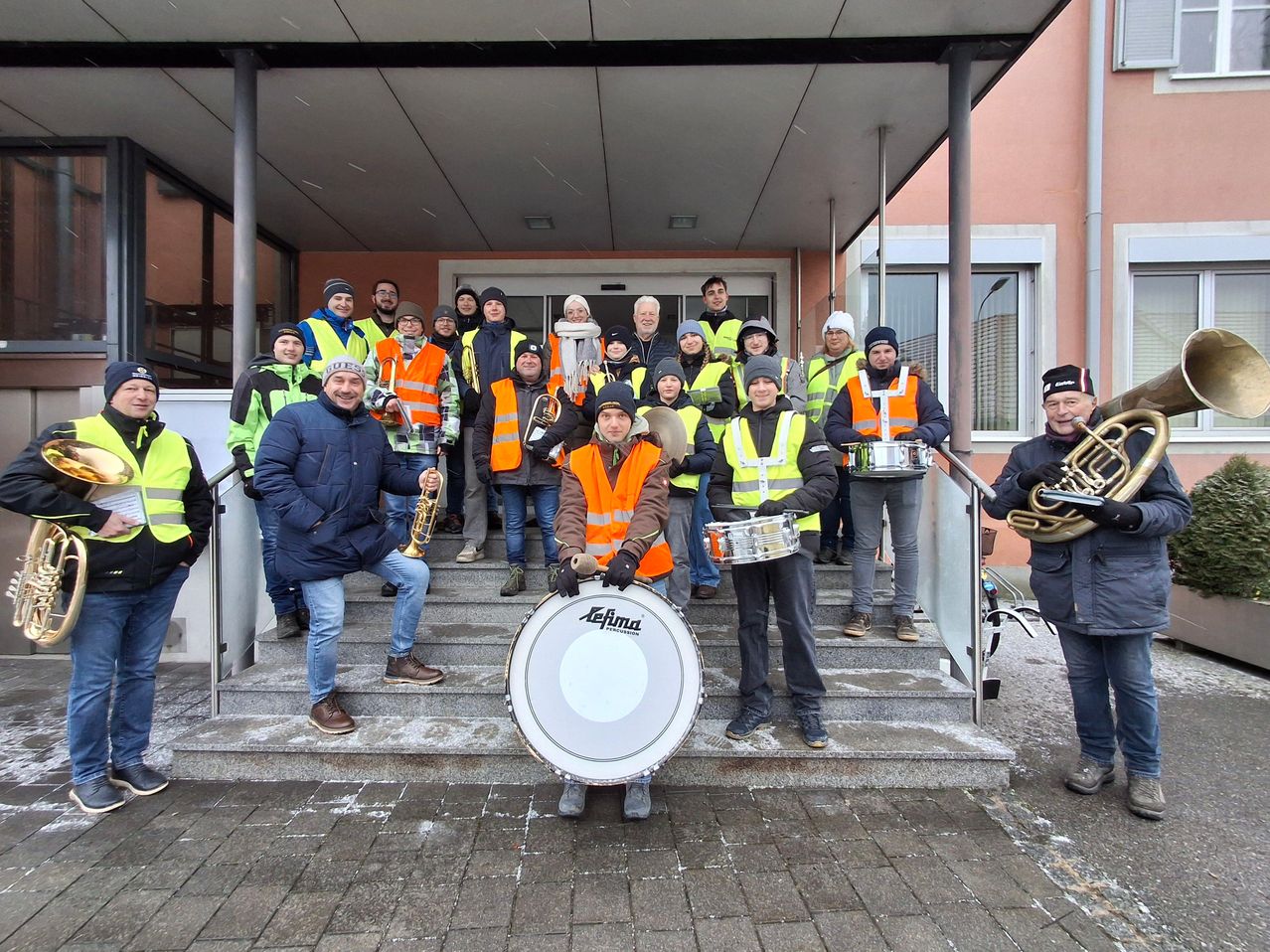 Eine Gruppe von Menschen in Warnwesten posiert auf einer Treppe vor einem Gebäude und hält Musikinstrumente in den Händen.