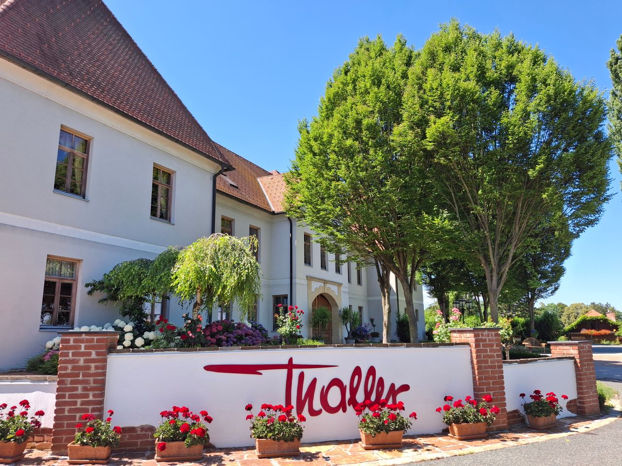 A white building with a red roof and a large sign that reads 'Thaller' in front of it. The building is surrounded by a garden with various flowers and trees.