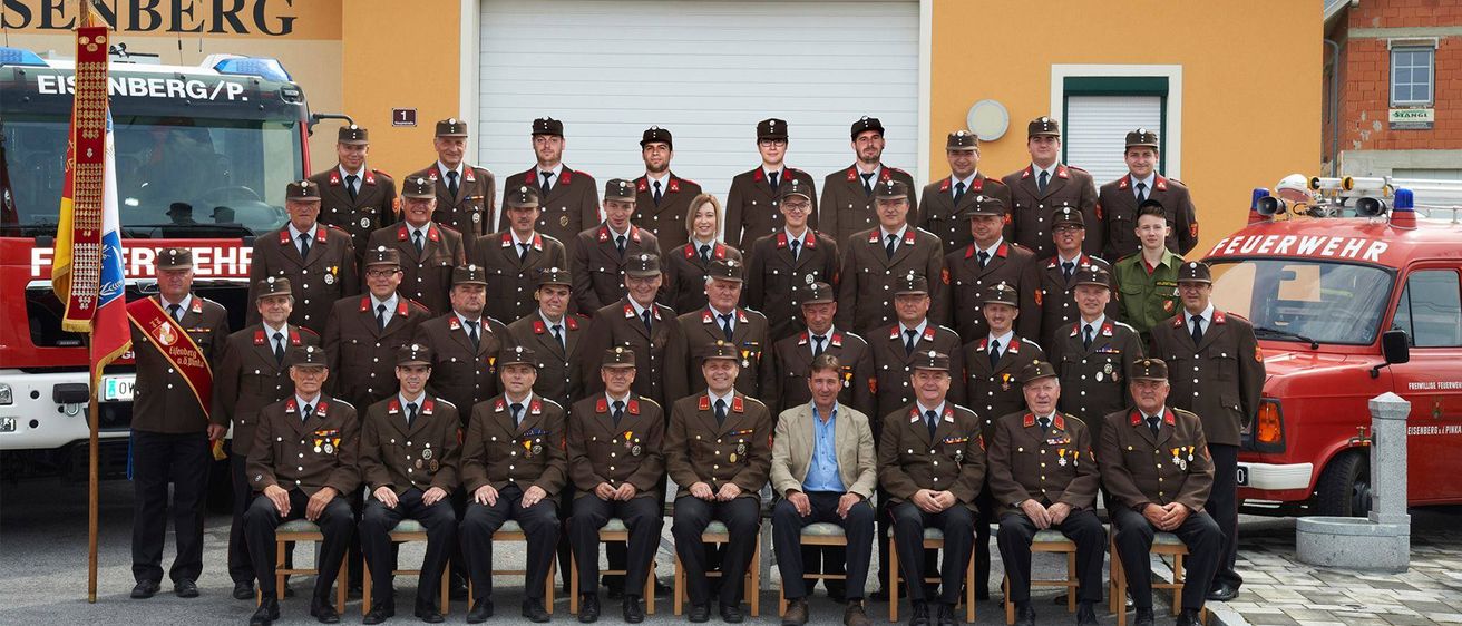 Eine Gruppe Feuerwehrleute in Uniform posiert für ein Foto vor einem Gebäude mit dem Wort 'Feuerwehr' darauf. Sie sitzen und stehen in Reihen.