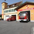 Zwei rote Feuerwehrfahrzeuge sind vor einem Feuerwehrhaus geparkt. Das Gebäude trägt die Aufschriften "Feuerwehr" und "Gemeindehaus Eisenberg".